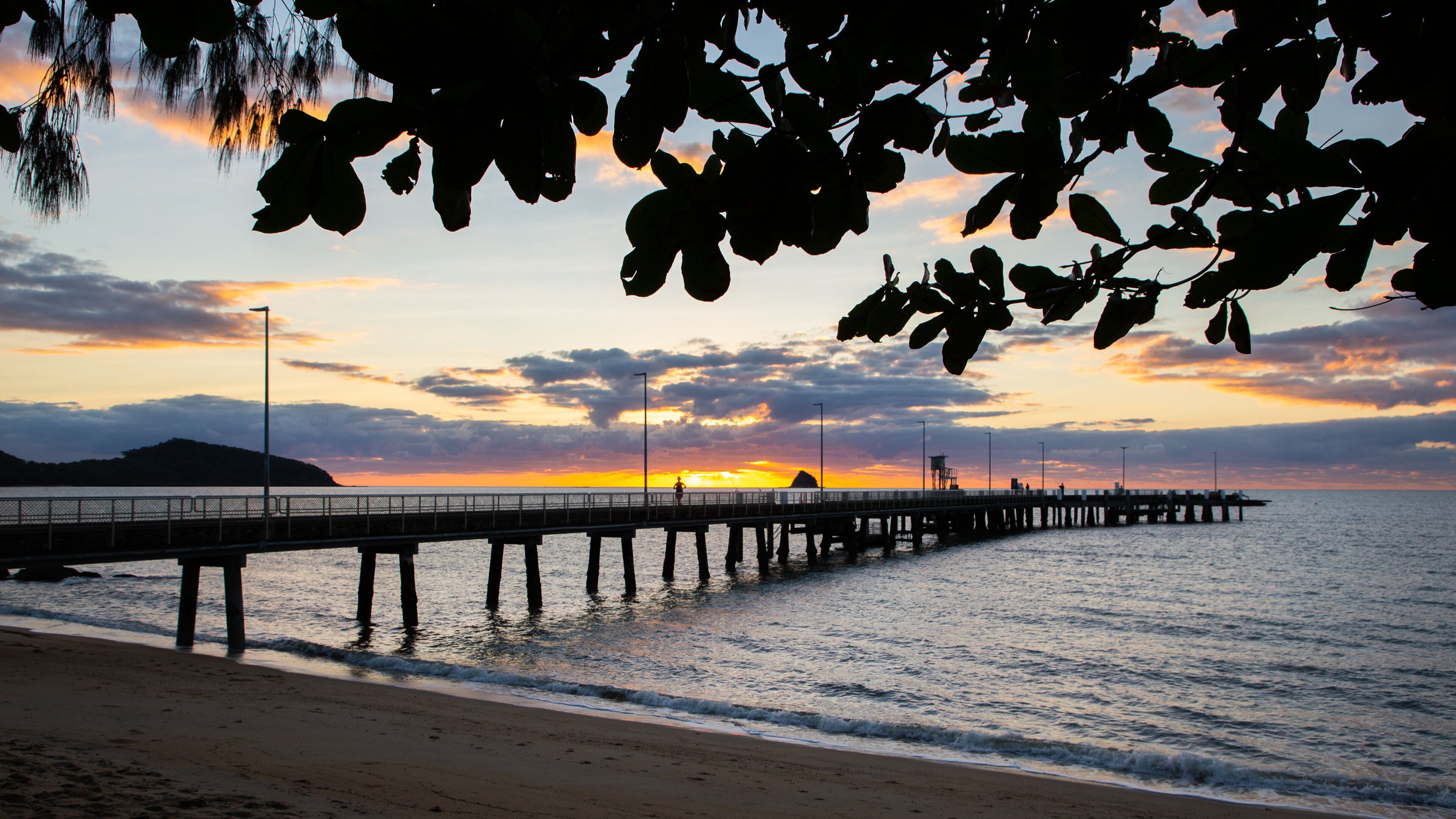 A pier extending into the water at sunset with a beach and mountains in the background.