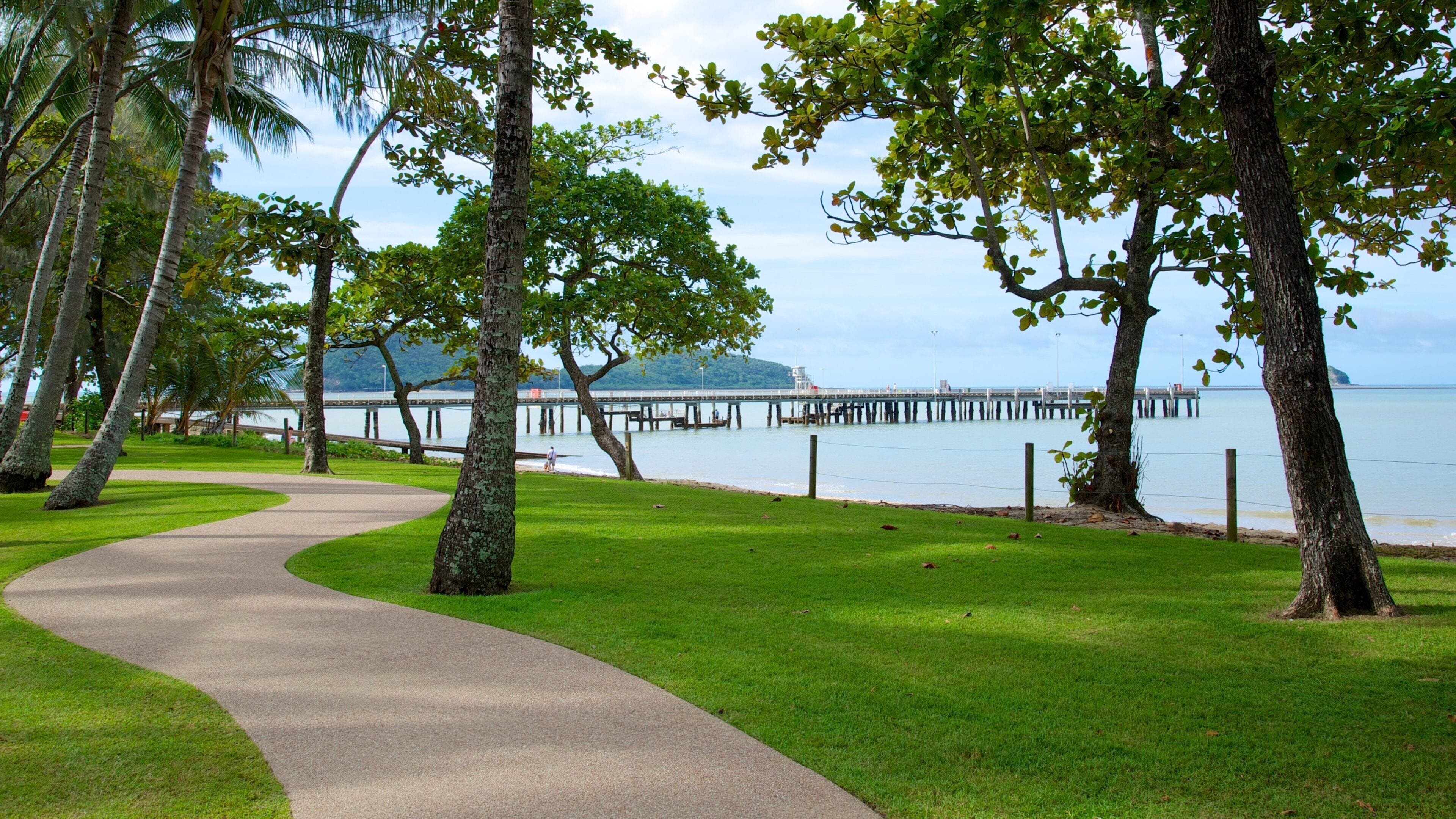 A paved path curving through a park with green grass and trees, leading to a body of water with a pier and a distant island.