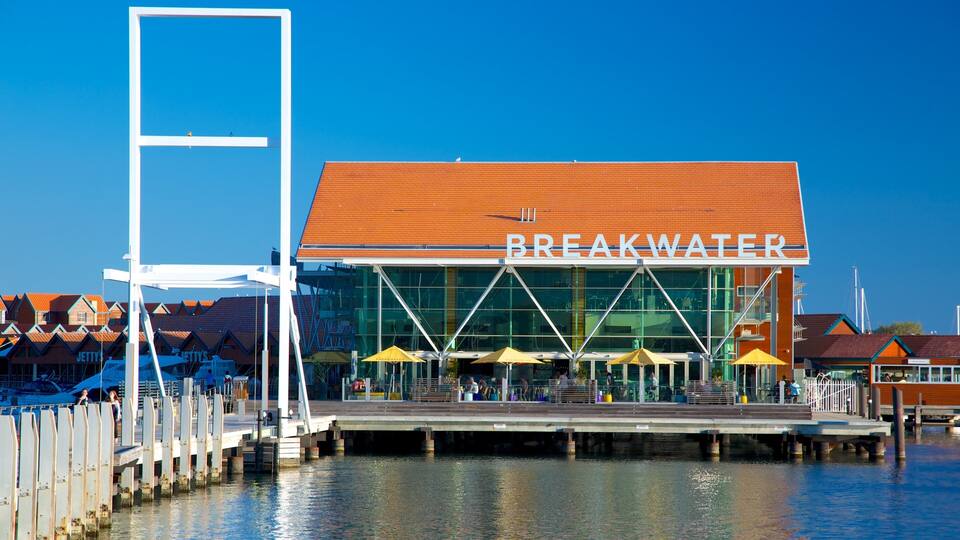 Un edificio frente al agua llamado "BREAKEWATER", con fachada de vidrio y techo rojo, rodeado por un muelle y sombrillas.