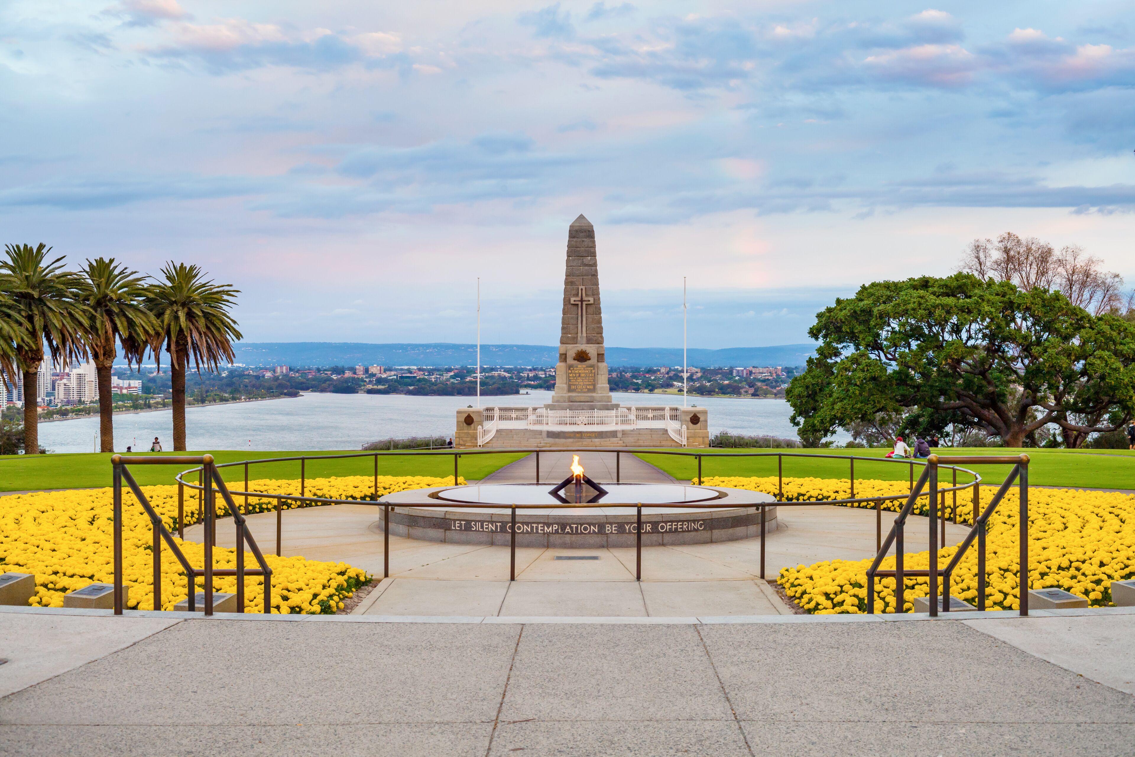 A monument with a cross, surrounded by yellow flowers and palm trees, overlooking a body of water.