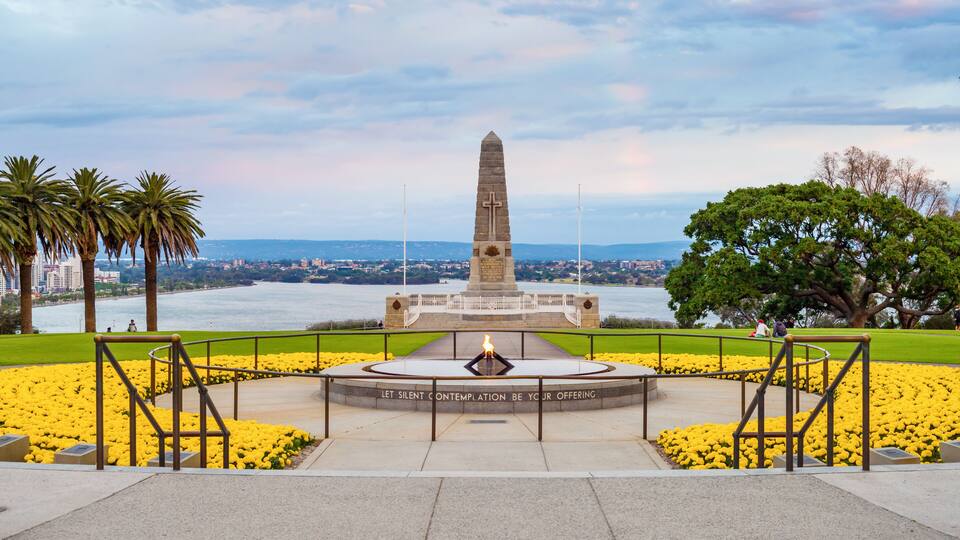 A monument with a cross, surrounded by yellow flowers and palm trees, overlooking a body of water.