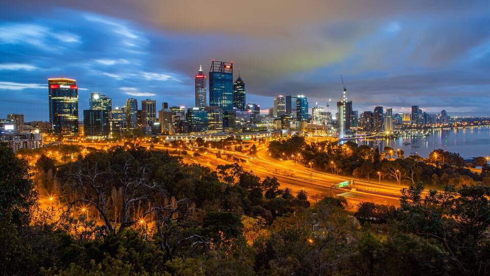 A city skyline at dusk with illuminated buildings and a river in the foreground.
