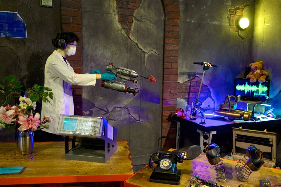 A person in a lab coat holding a toy gun, a computer with a screen, and various electronic devices on a table.