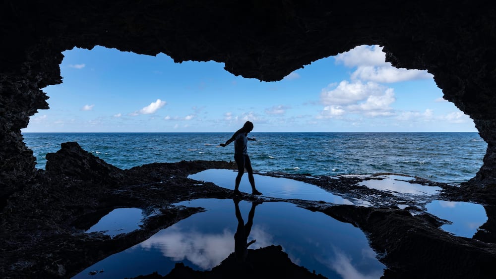 Une personne se tient dans une grotte, d’où l’on aperçoit l’océan.