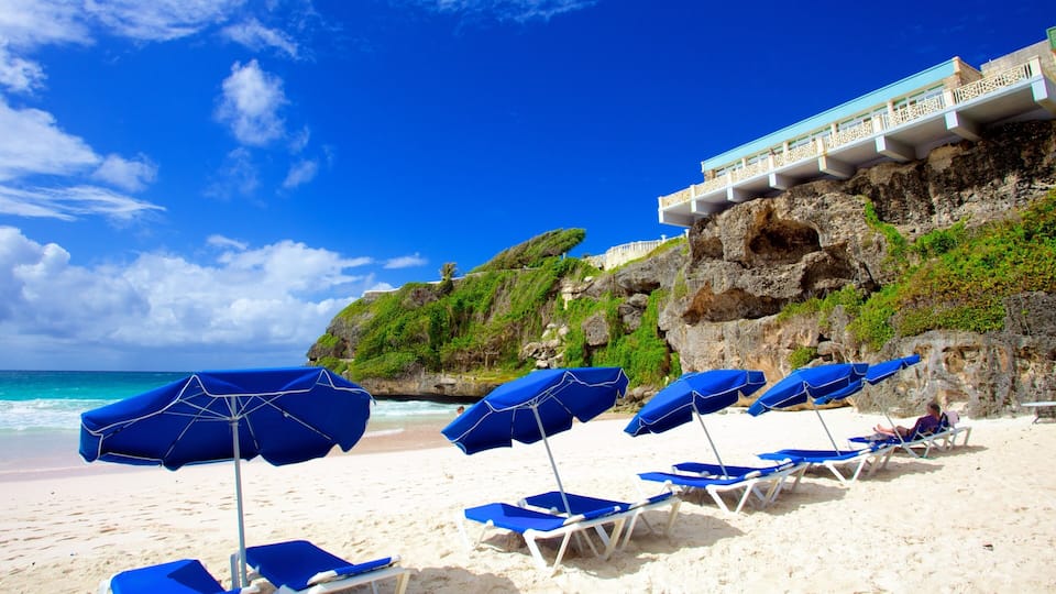 A beach with blue umbrellas and lounge chairs, a rocky cliff with greenery, and a building with balconies overlooking the ocean.