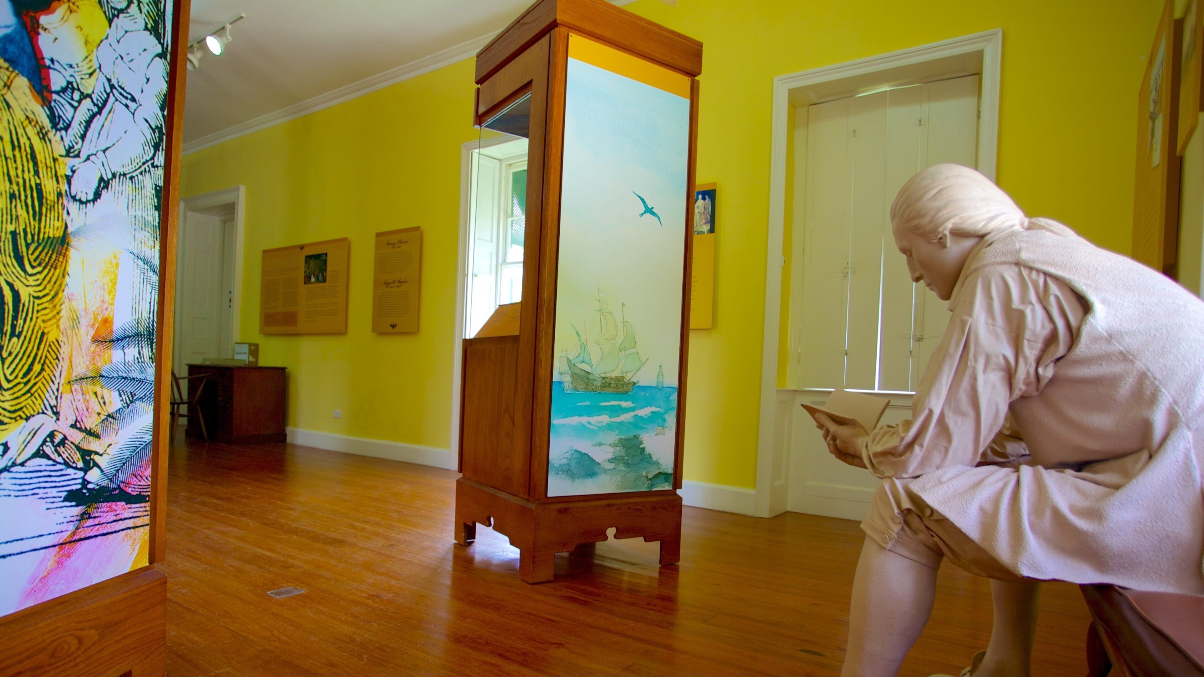 A person reading in a room with yellow walls, wooden floors, and a large painting of a ship on a stand.