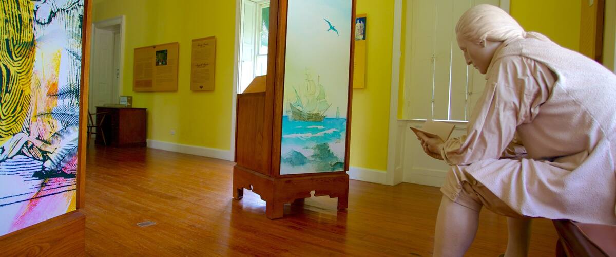 A person reading in a room with yellow walls, wooden floors, and a large painting of a ship on a stand.