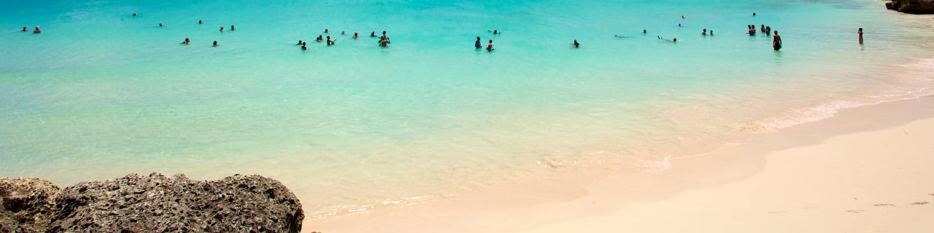 Une plage aux eaux turquoise et limpides, où l’on voit des gens se baigner et des bateaux au loin.