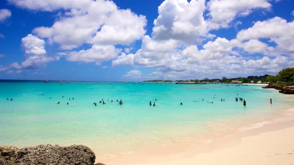 A turquoise beach with clear waters, people swimming, and boats in the distance.