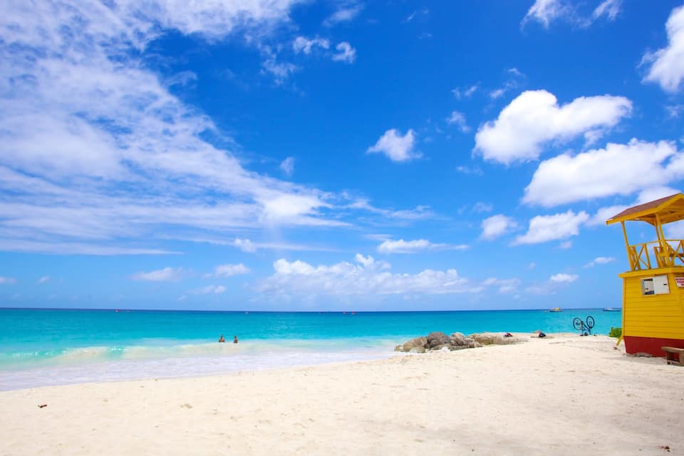 Uma praia com águas turquesa cristalinas, uma torre de salva-vidas amarela e um céu azul límpido com algumas nuvens.