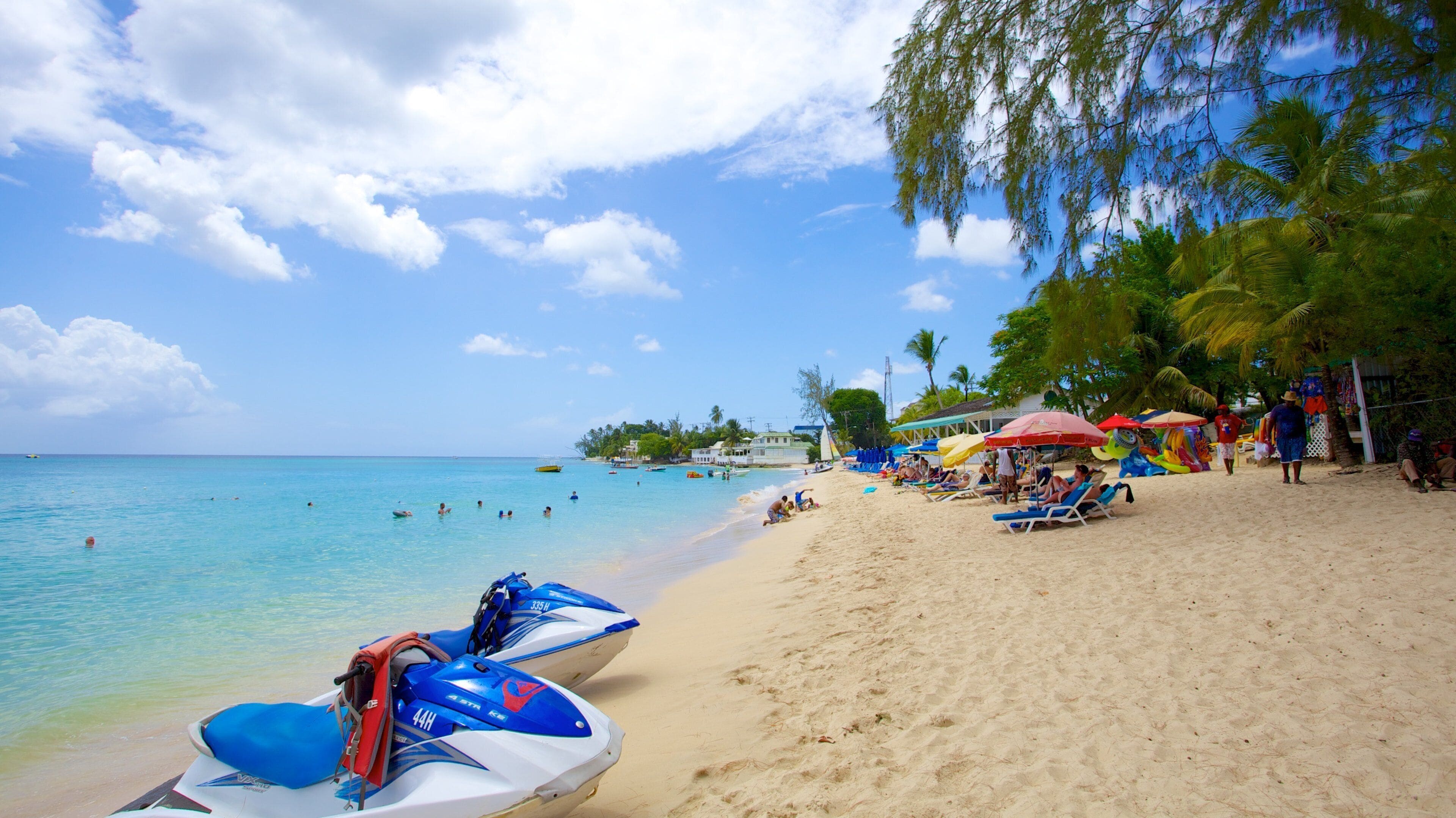 A beach with clear turquoise water, a sandy shore, and a few jet skis parked on the sand.