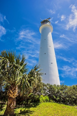 Un grand phare blanc doté d’un balcon, entouré d’une végétation luxuriante et de palmiers, se dressant sur un ciel bleu dégagé.