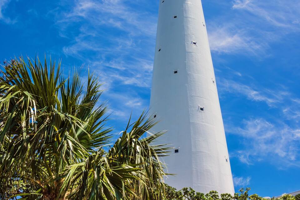 Un alto faro blanco con un balcón, rodeado de exuberante vegetación y palmeras bajo un cielo azul despejado.