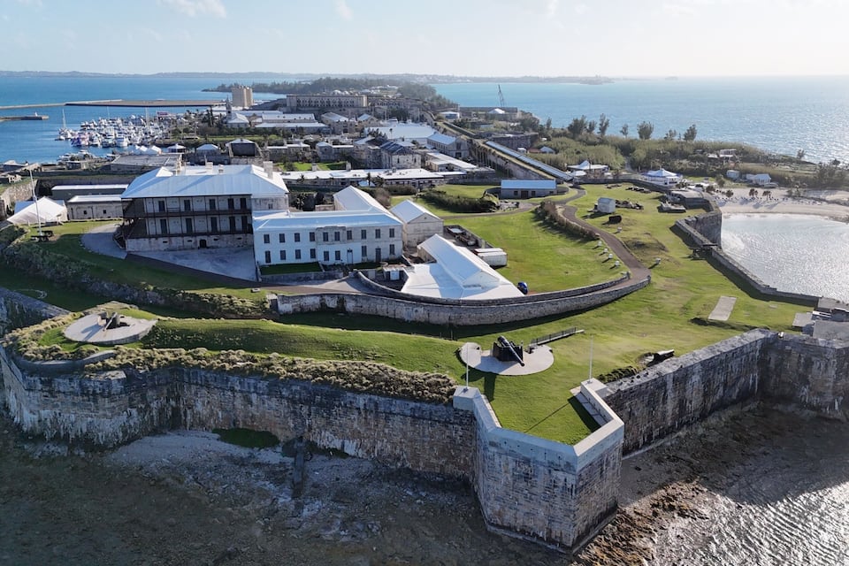 An aerial view of a historic fort with a large white building, a surrounding moat, and a harbor with boats.