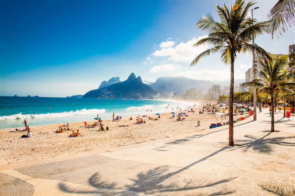 Una playa con palmeras, olas del océano y montañas al fondo.