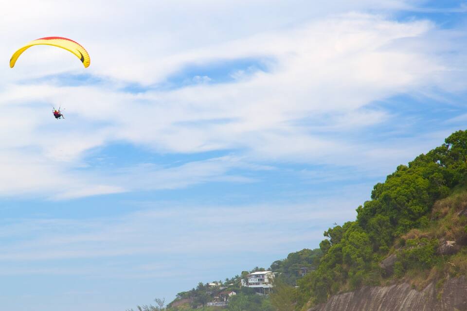 A person paragliding over a coastal area with a cliff and houses in the background.