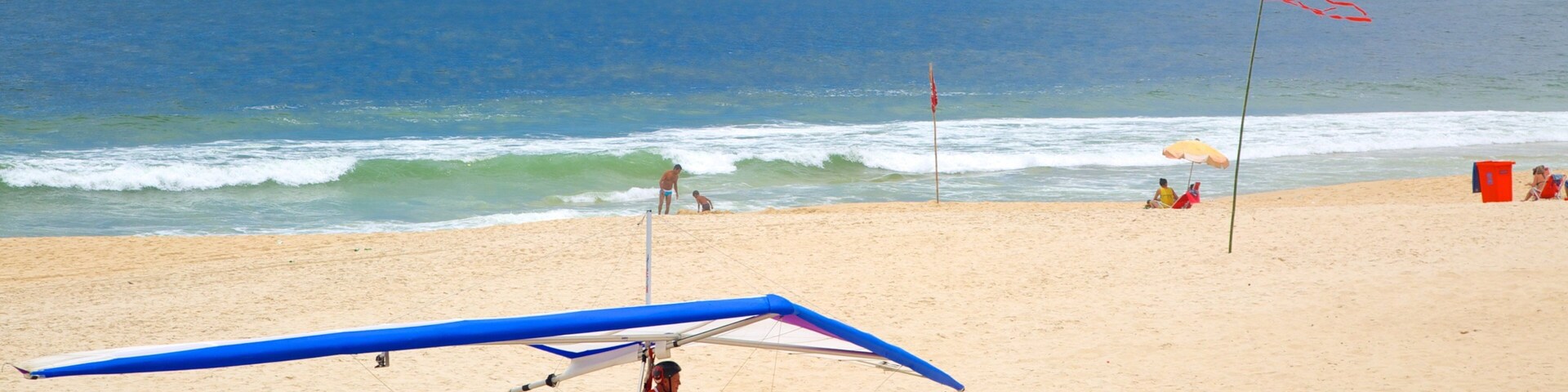 Uma praia com um paraquedas azul, pessoas caminhando e uma bandeira.