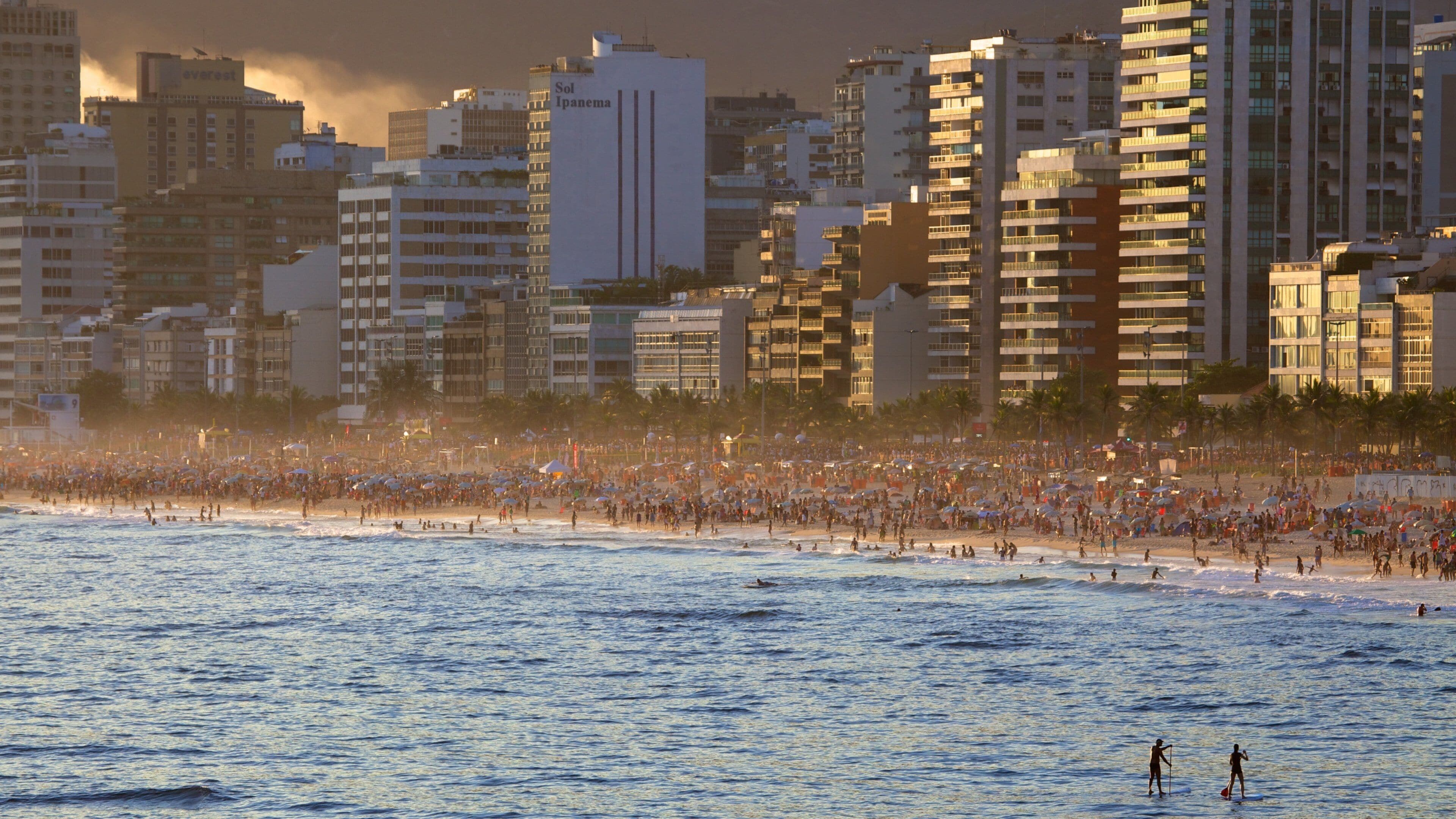 A crowded beach with people and umbrellas, and tall buildings in the background.
