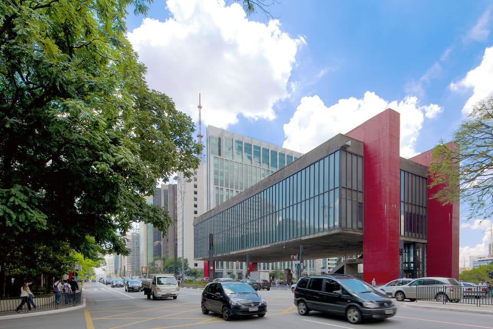 Un bâtiment moderne doté de grandes fenêtres en verre et d’éléments décoratifs rouges, entouré d’arbres et bordé de rues animées.