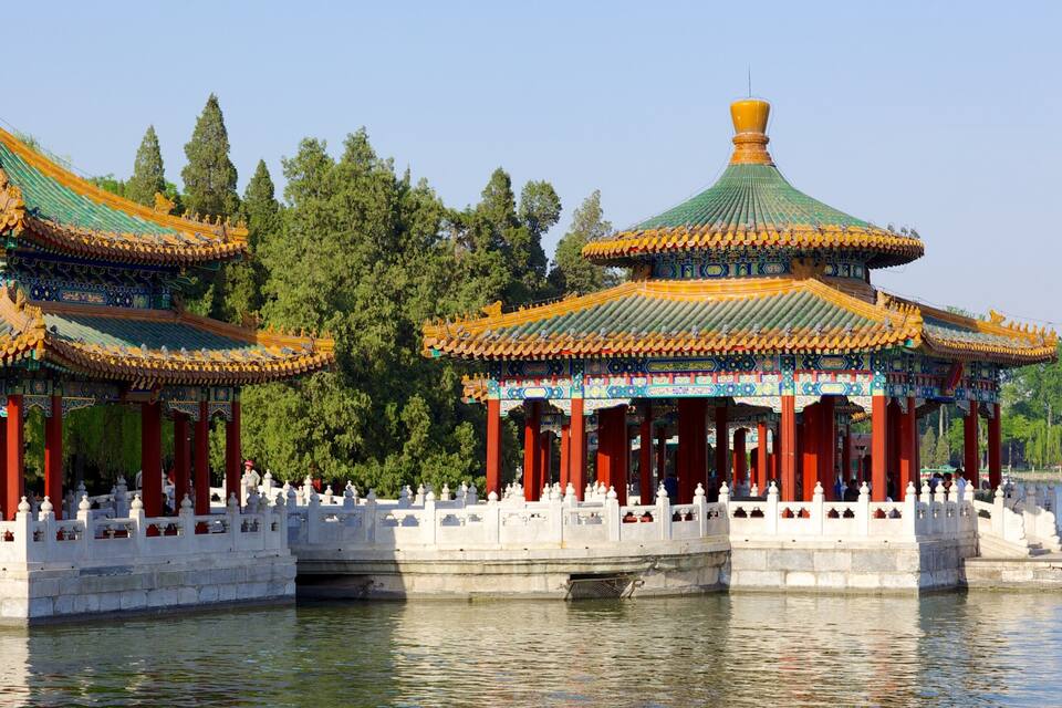 Een traditioneel Chinees paviljoen met versierde daken en rode zuilen, gelegen op een stenen brug over een rustig waterlichaam.