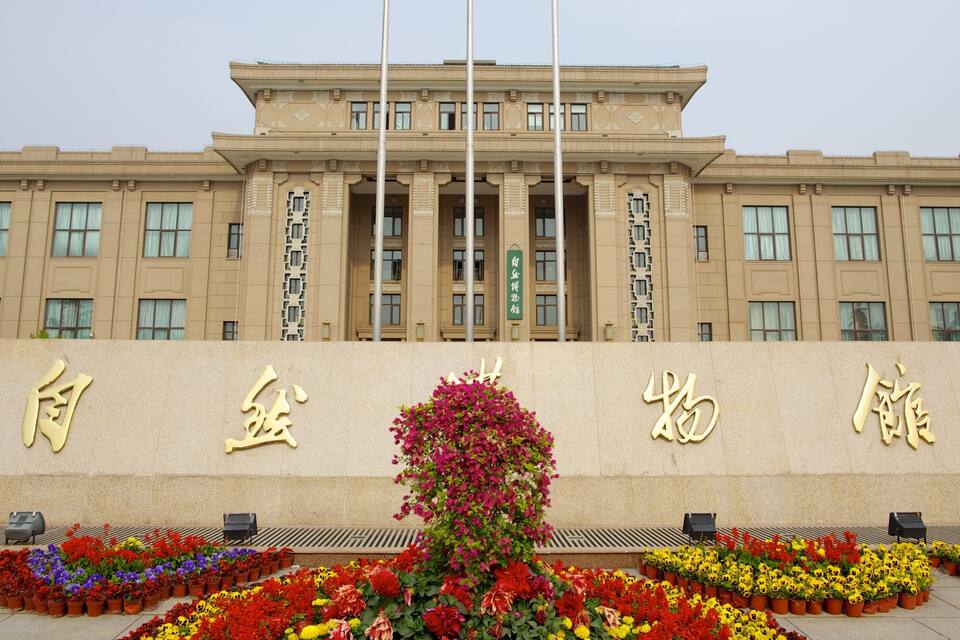 A large building with columns and a sign in front. There are colorful flowers in front of the building.