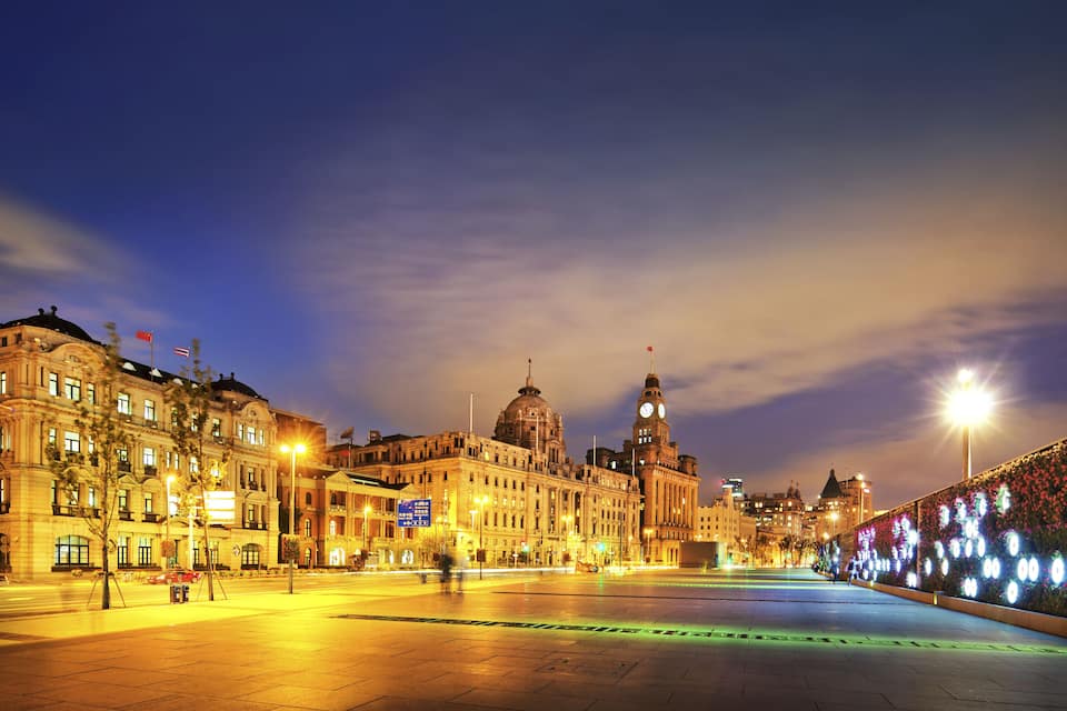 Uma praça histórica ao entardecer, com edifícios iluminados e uma torre do relógio imponente.