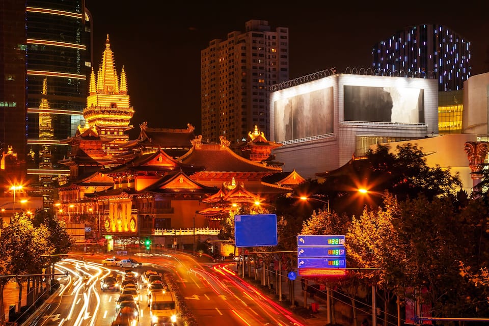 A night view of a city with traditional architecture, modern buildings, and a busy road with light trails.