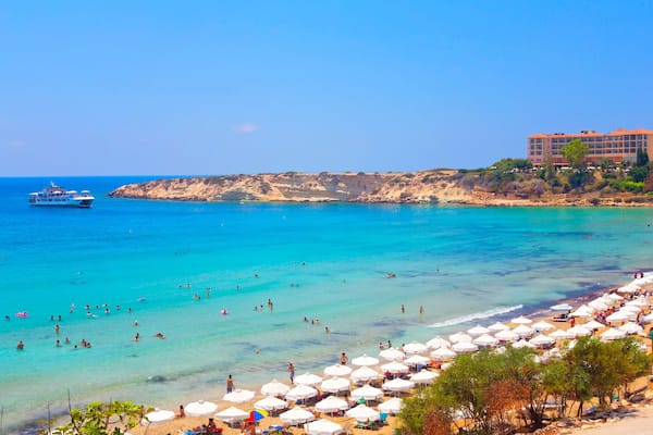 Une plage à l’eau turquoise et limpide, un navire de croisière et un bâtiment aux toits rouges.