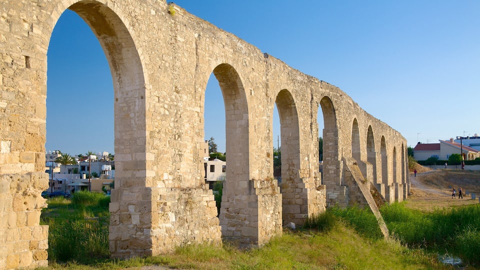An ancient stone aqueduct with multiple arches, surrounded by grassy areas and residential buildings in the background.