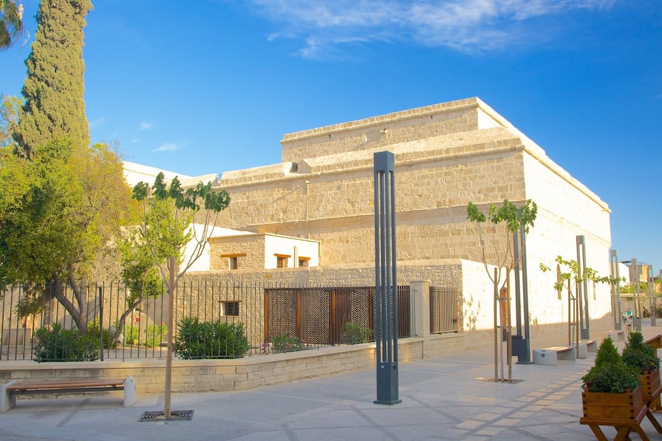 A historic stone building with a flat roof, surrounded by trees and a paved walkway.