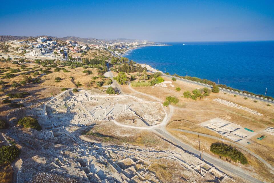 An aerial view of a coastal archaeological site with ruins, a road, and a beach.