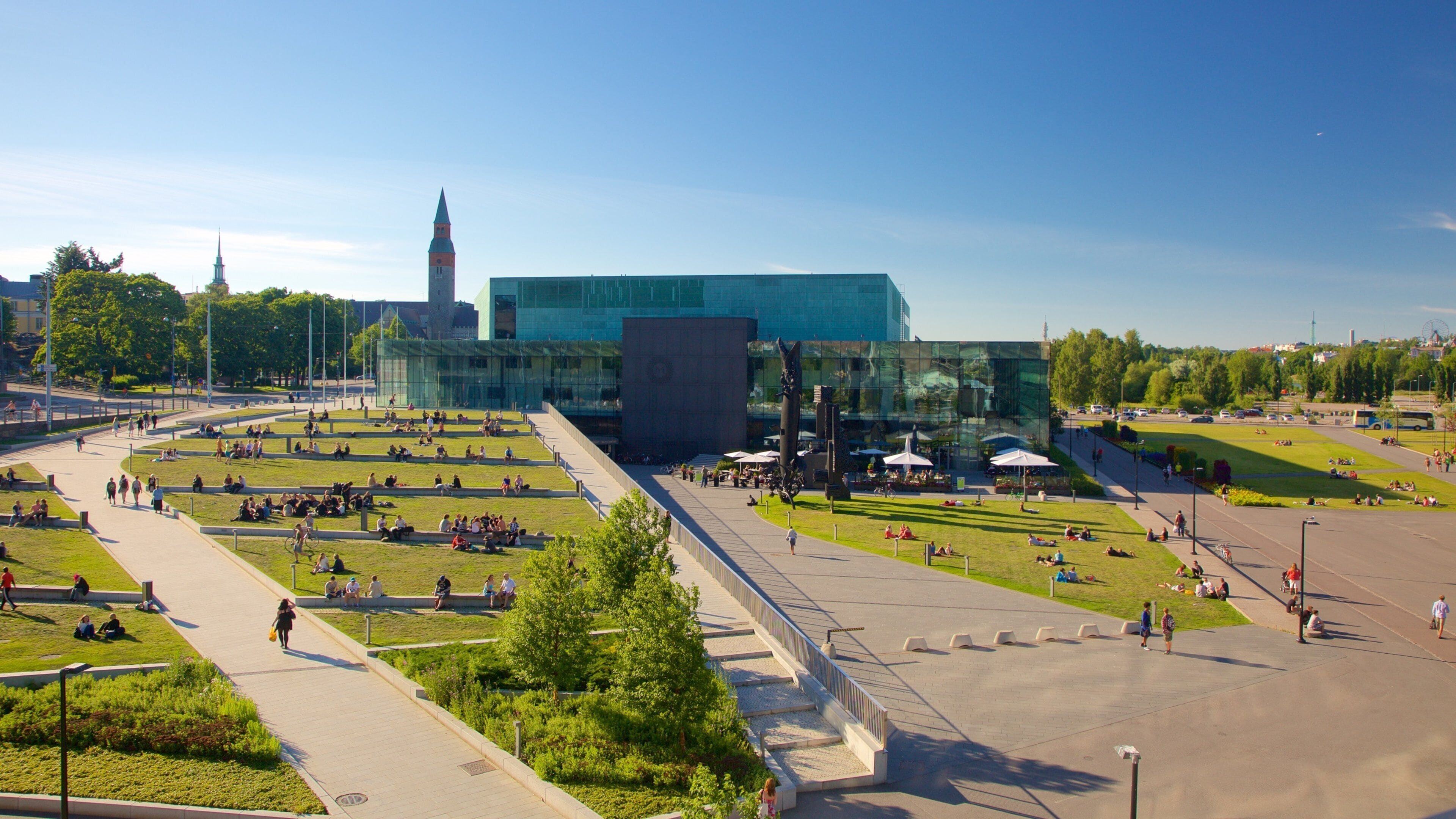 A modern plaza with people sitting on grass and walking paths, surrounded by green spaces and a large glass building with a tower.