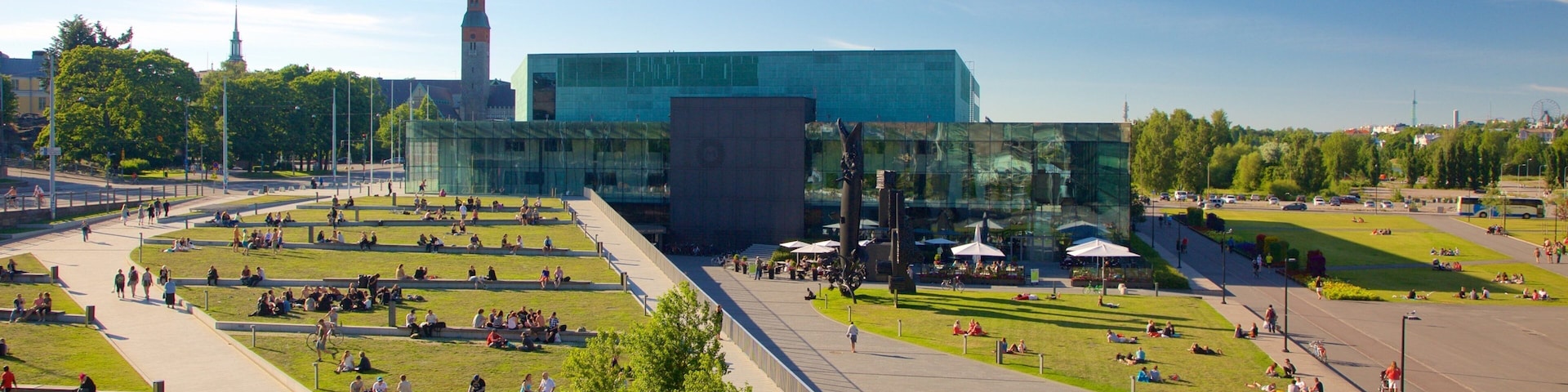 Una plaza moderna con gente sentada sobre el césped y caminos peatonales, rodeada de áreas verdes y un gran edificio de cristal con una torre.