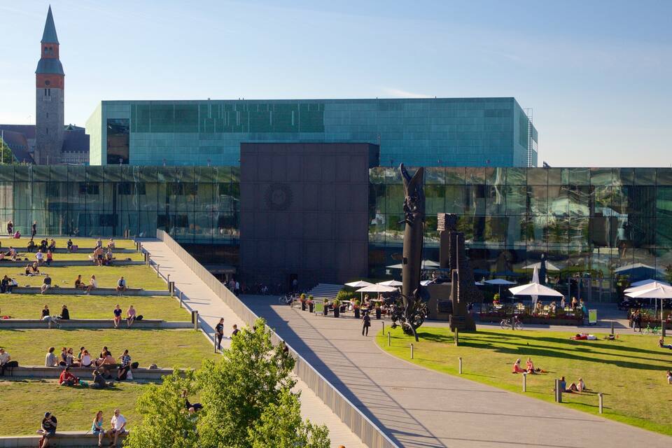 En moderne bygning med glasfacade, en central gårdsplads med siddeområder, og et historisk tårn i baggrunden.
