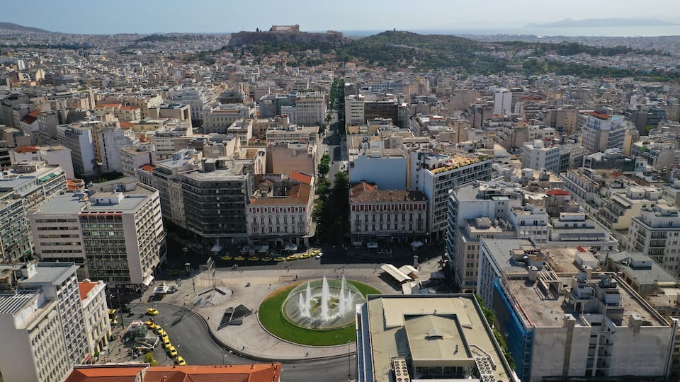 A cityscape with a prominent roundabout, historic buildings, and a distant view of a hill with ruins.