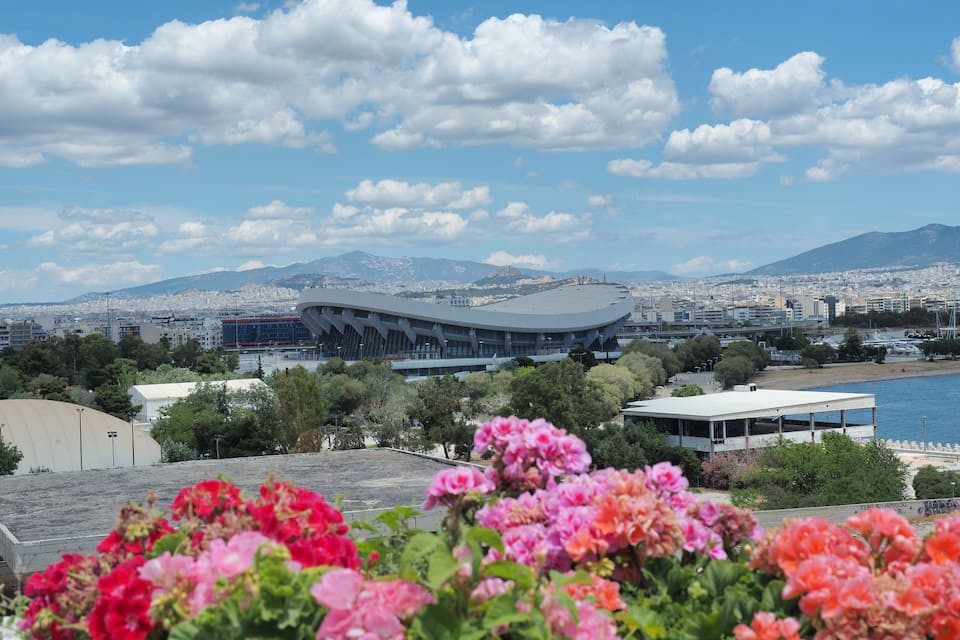 Een modern stadion met een uniek dakontwerp, omgeven door een stadsgezicht en bergen op de achtergrond.