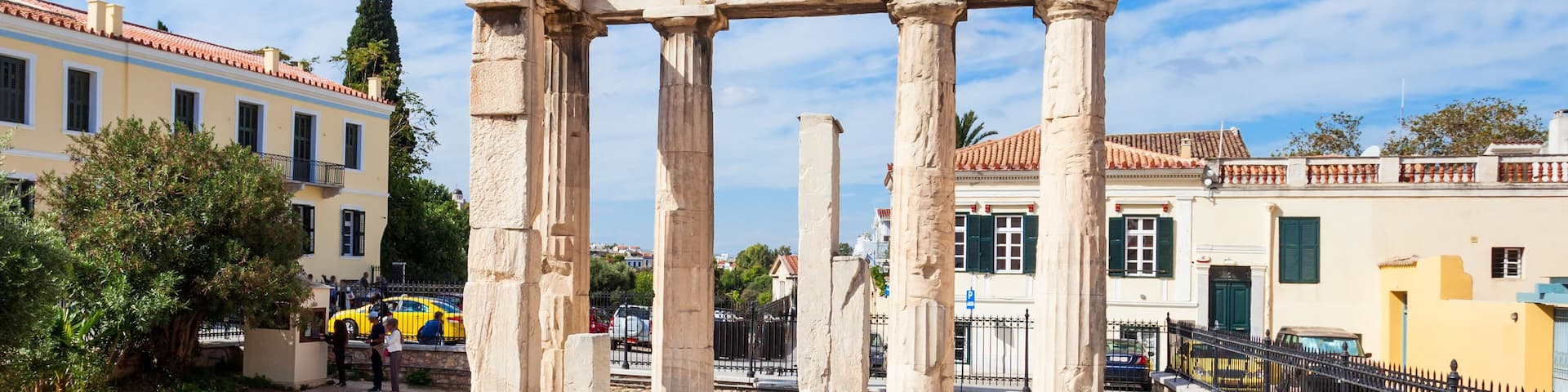 Ancient stone ruins with columns and a pediment, surrounded by modern buildings and a clear blue sky.