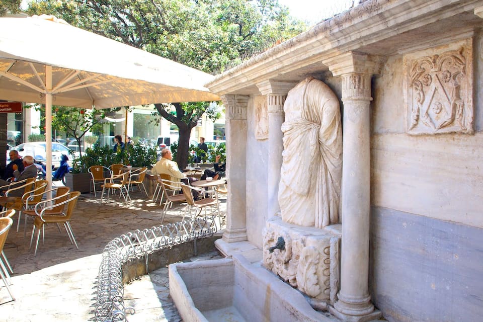 Ein klassisches Bauwerk mit Säulen und aufwendigen Schnitzereien, umgeben von Außenplätzen und einem Brunnen.