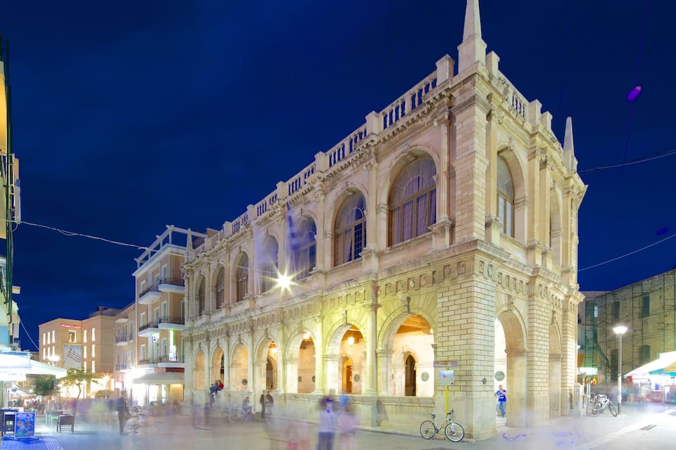 A historic building with ornate architecture, a large open plaza, and surrounding modern buildings.