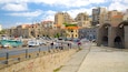 A coastal cityscape with historic buildings, a harbor with boats, and a stone wall with graffiti.