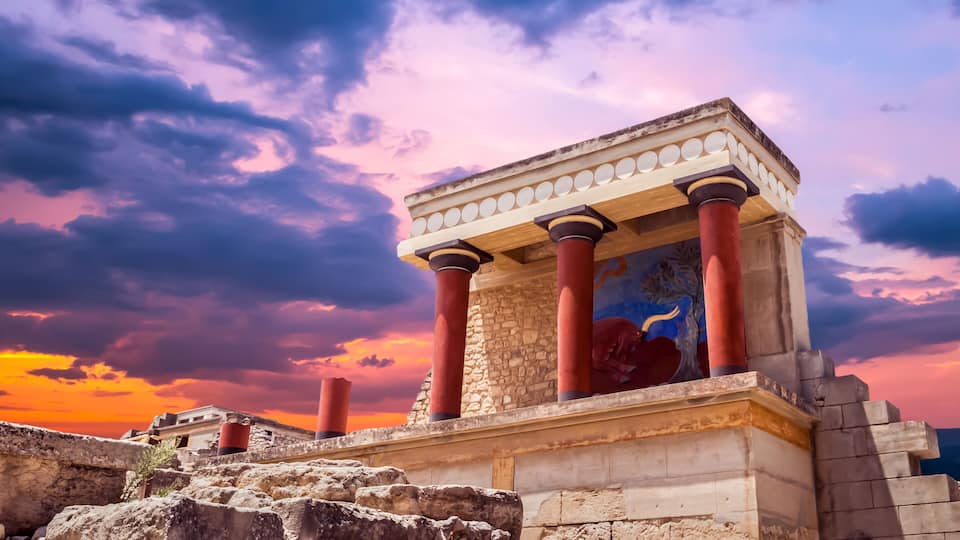 An ancient temple with red columns and a stone base, set against a dramatic sunset sky.