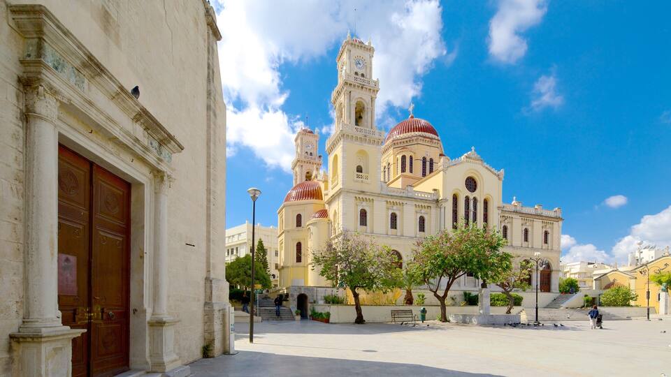 A historic church with a clock tower and domes, surrounded by a plaza with trees and benches.
