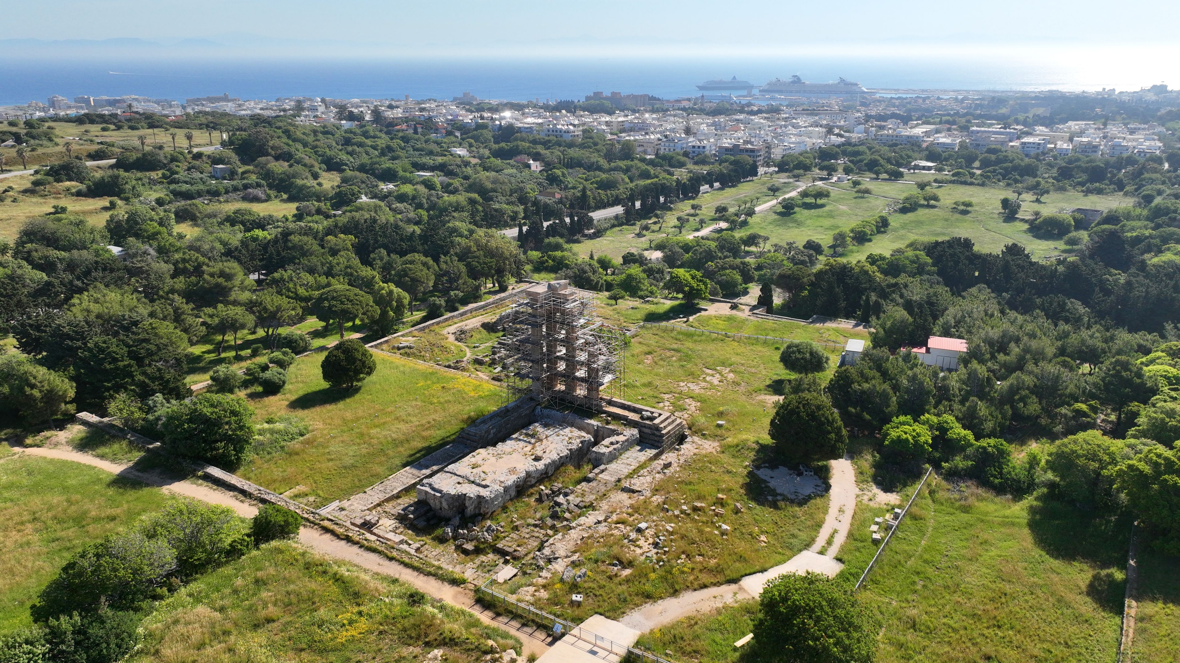 An ancient archaeological site with ruins and a large tree in the center, surrounded by lush greenery and a coastal cityscape in the background.