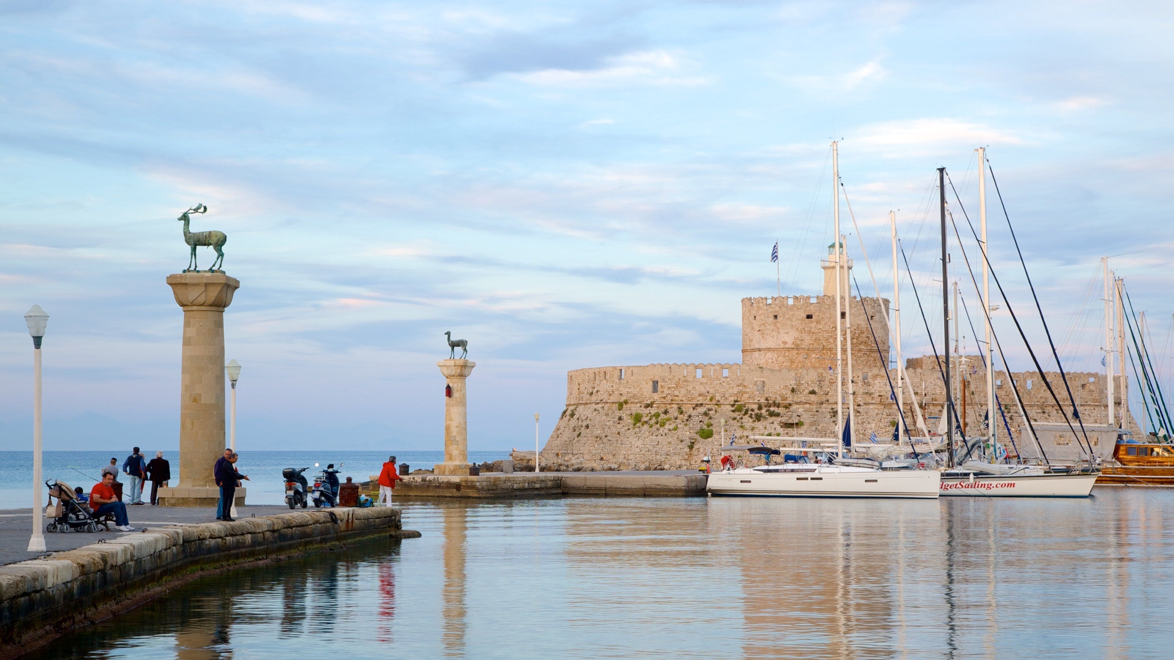 A coastal scene with a historic stone castle, two stone pillars with animal statues, a calm sea, and several sailboats docked near the shore.