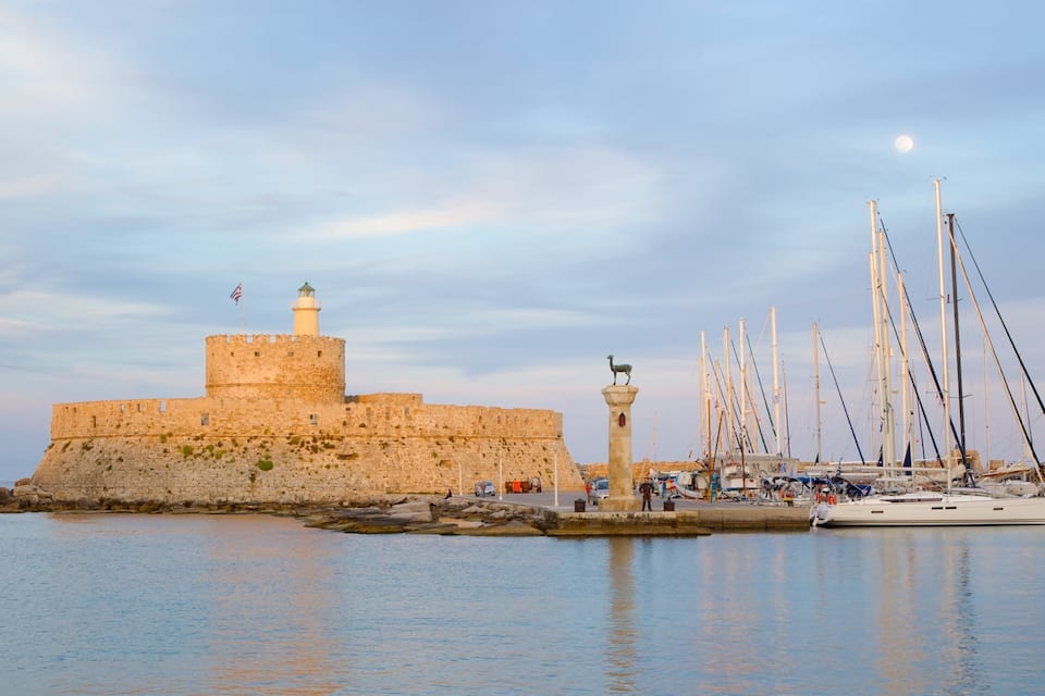 Une forteresse historique abritant un phare, un quai où sont amarrés des voiliers et une mer calme.