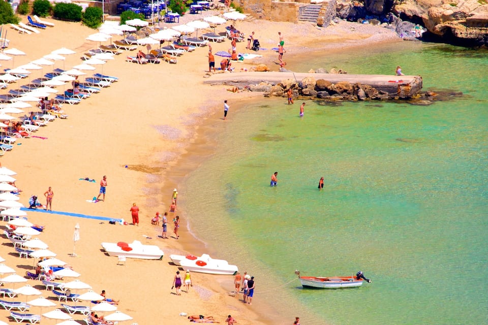 A beach with rows of sun loungers and umbrellas, clear turquoise water, and a rocky breakwater.