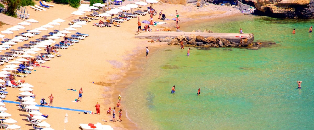 A beach with rows of sun loungers and umbrellas, clear turquoise water, and a rocky breakwater.