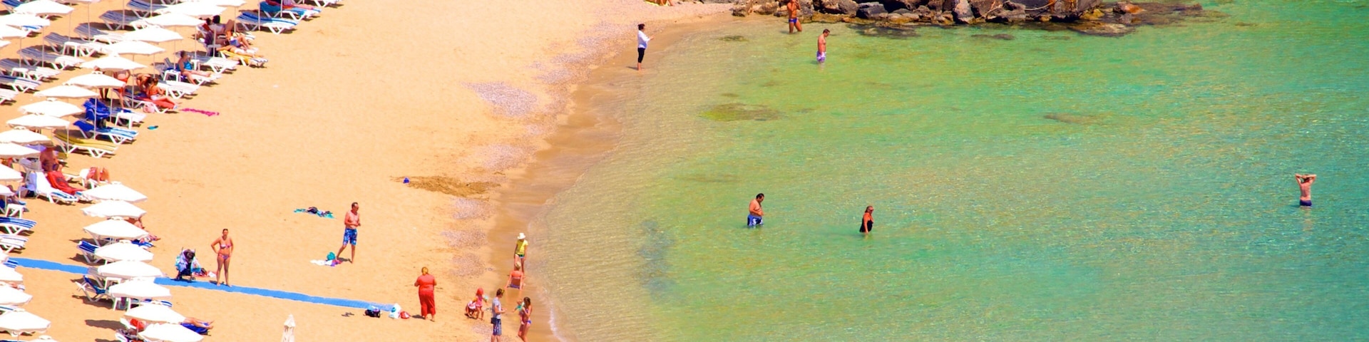 A beach with rows of sun loungers and umbrellas, clear turquoise water, and a rocky breakwater.