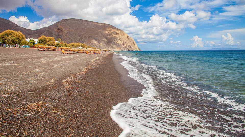 A pebbly beach with waves, a mountain in the background, and a clear sky.