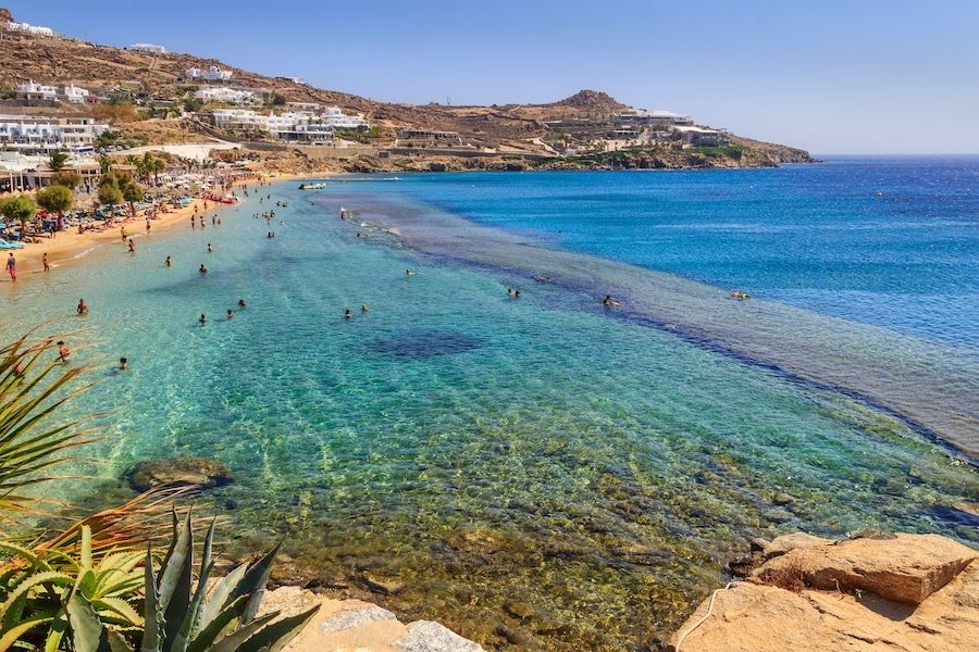 Una playa costera con aguas turquesas cristalinas, costa rocosa y edificios blancos en una colina.